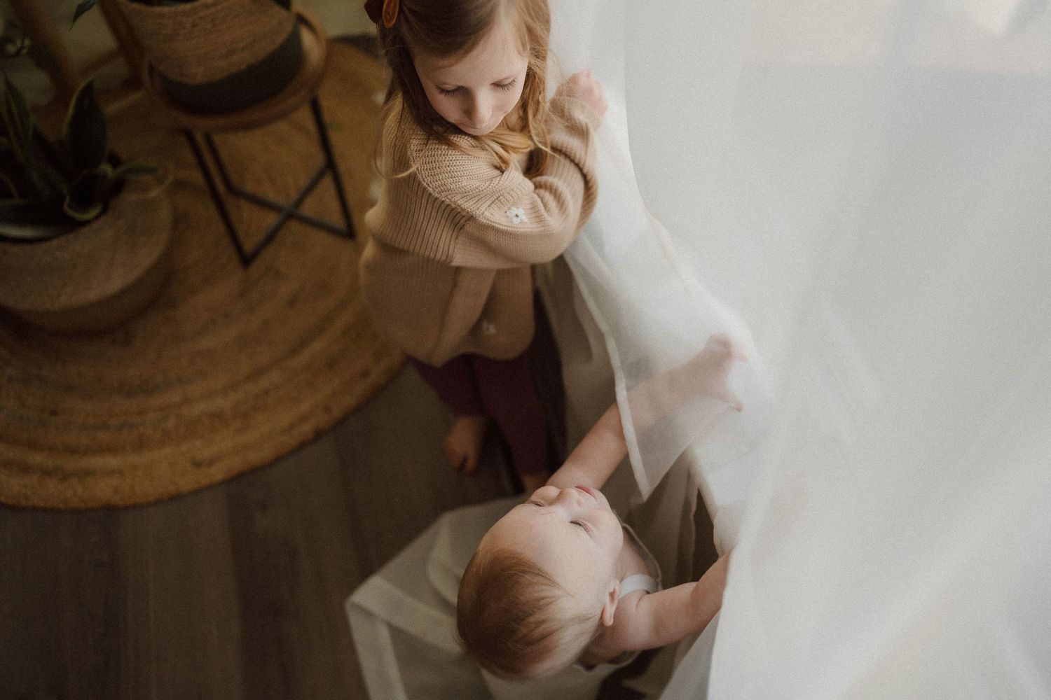 Two children playing with white curtains in a cozy home setting.