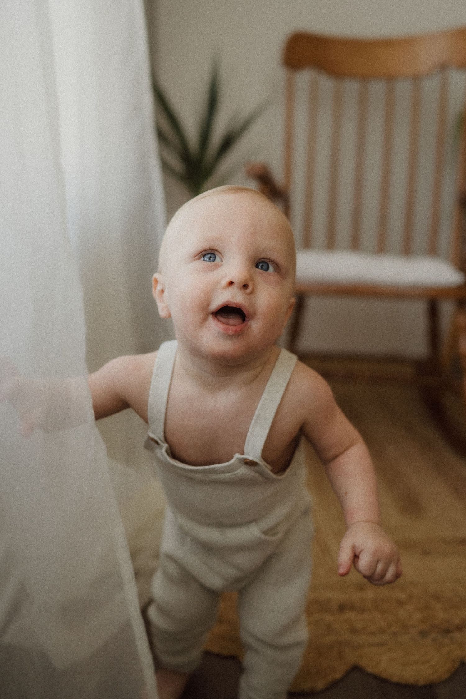 Excited baby in neutral-colored romper standing by flowing white curtains.