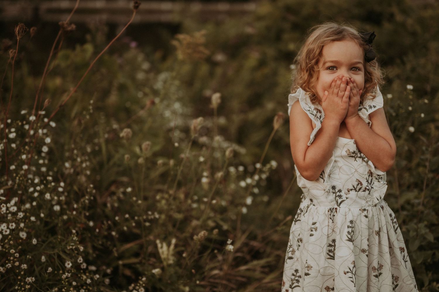 Child in a white floral dress laughing with hands covering face in a meadow.