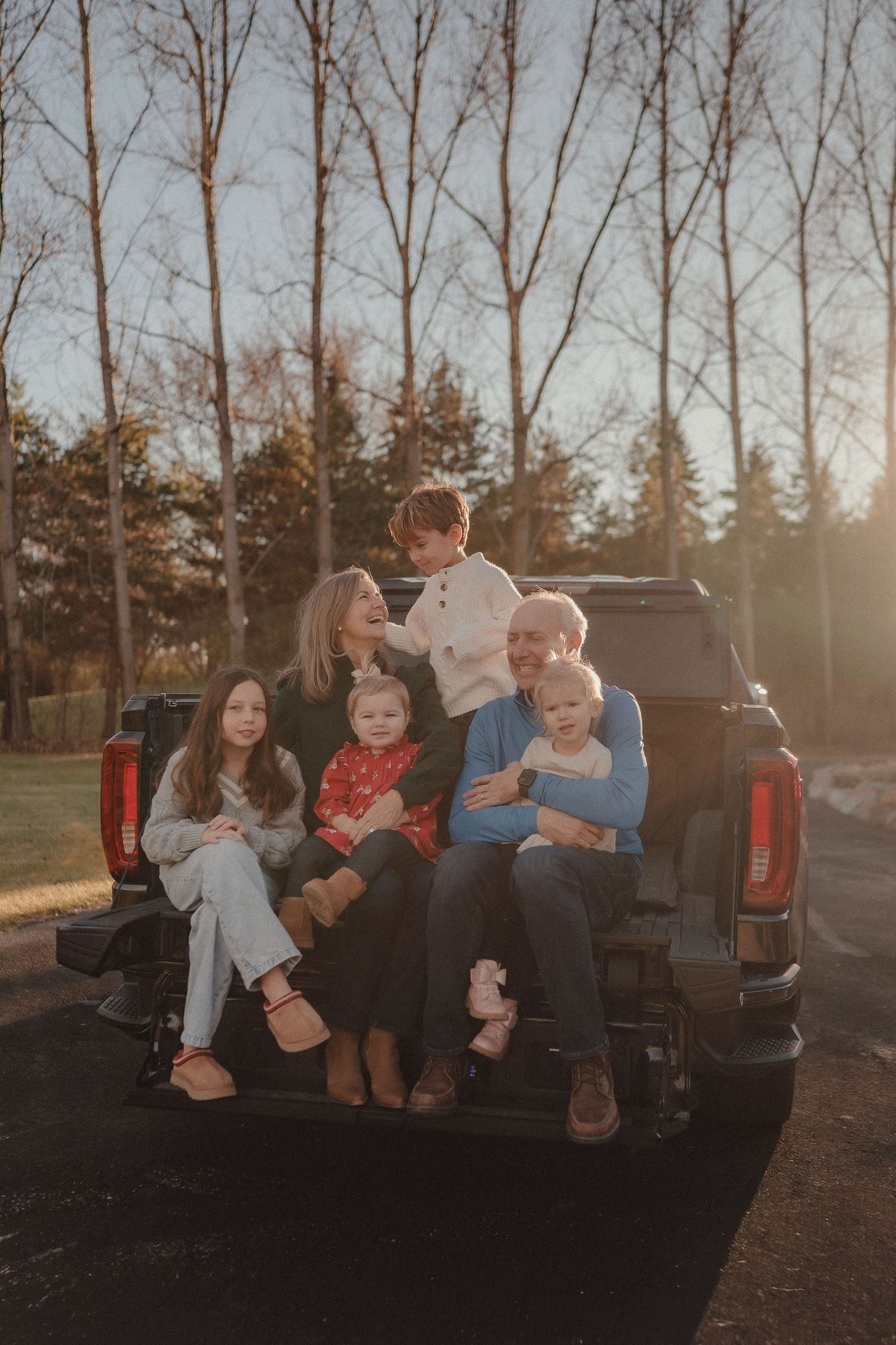 A family sits together in the back of a pickup truck at sunset with bare trees silhouetted against the sky.