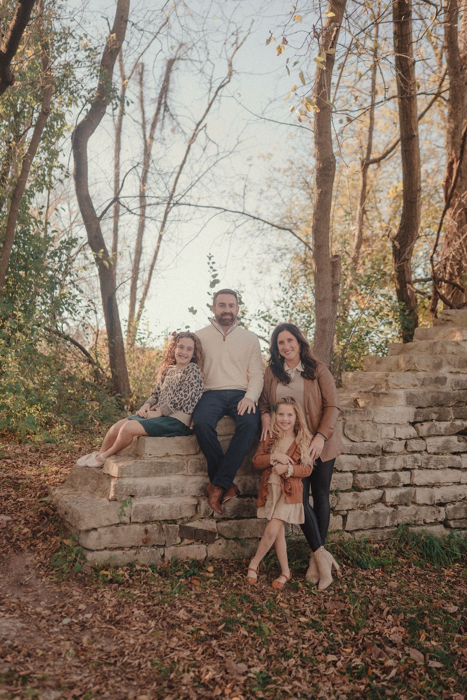 Family sitting together on rustic stone wall surrounded by autumn trees during golden hour outdoor photoshoot.