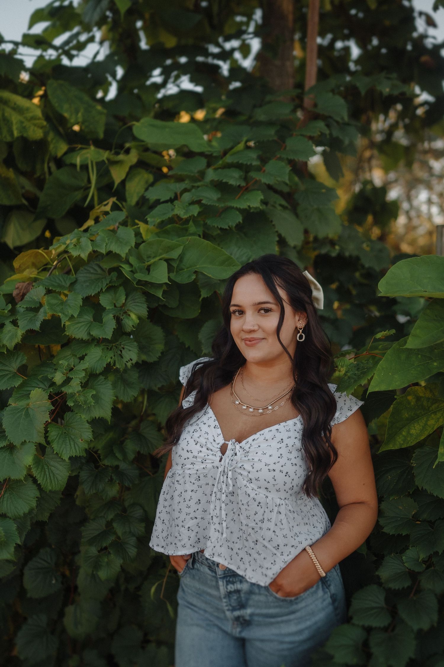 Portrait against lush green foliage backdrop wearing white floral top and jeans.