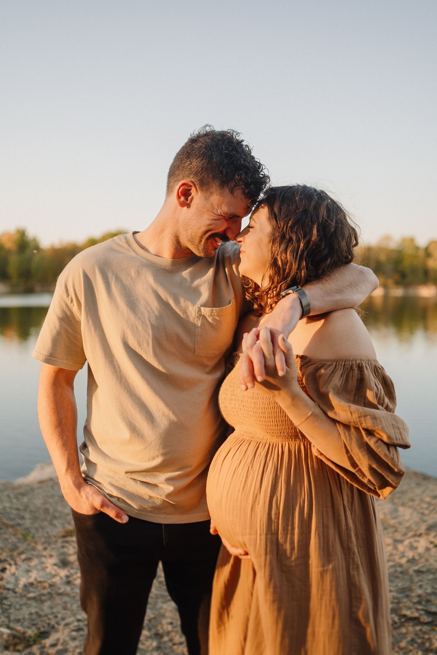 Intimate maternity portrait of couple sharing tender moment by lake at sunset.