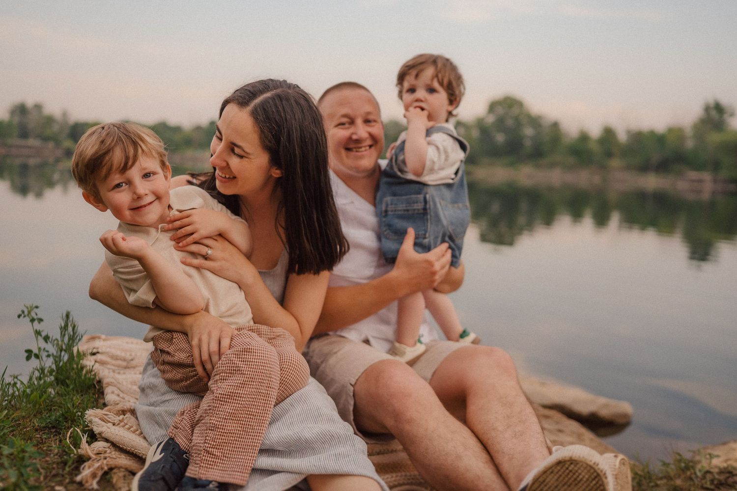 Family shares tender moments together while relaxing on rocks beside a serene lake.