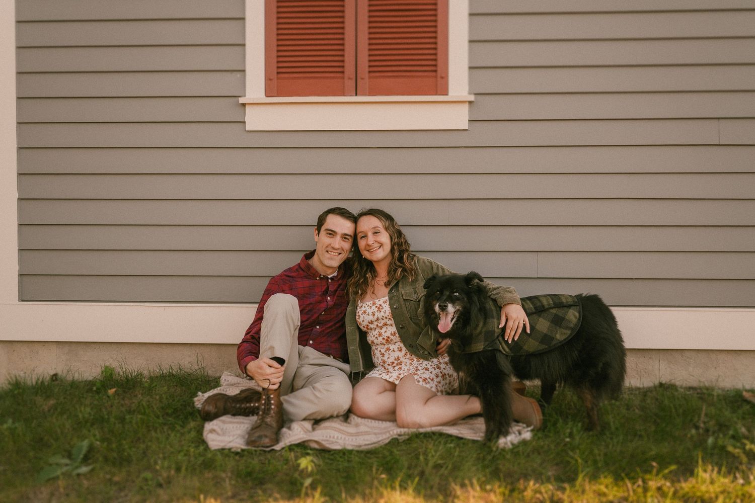 A couple sits with their black dog outside a grey house at sunset.