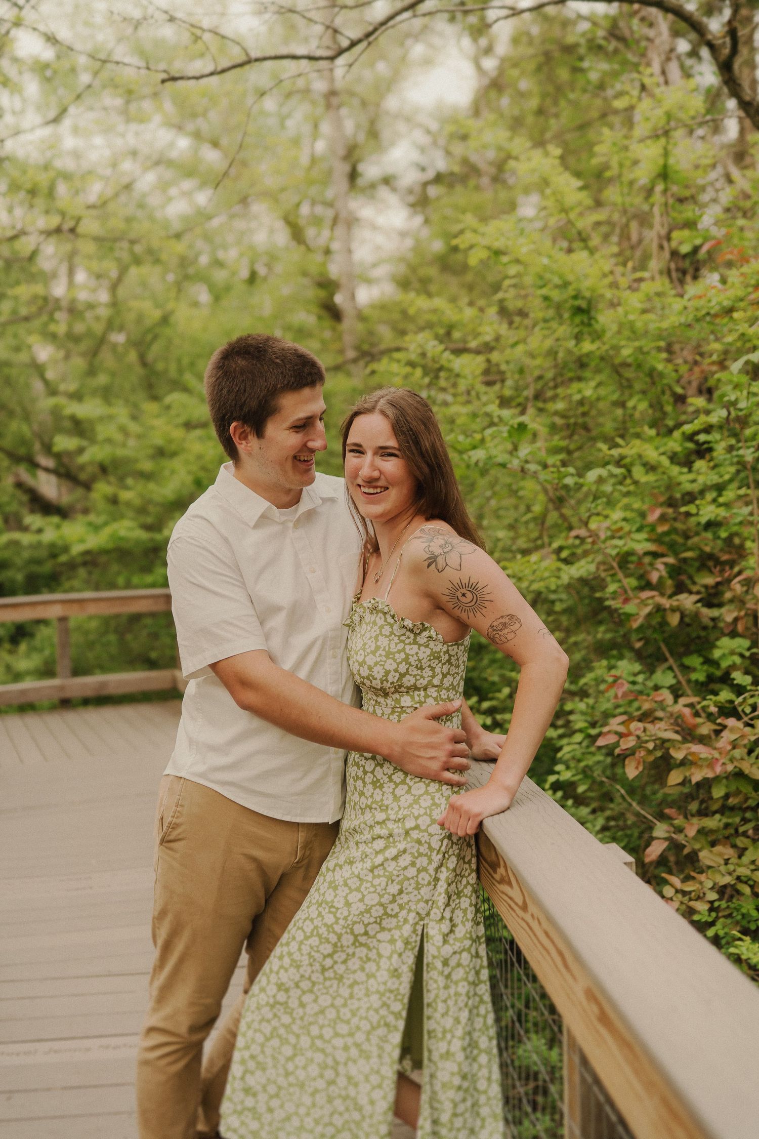 Romantic couple laughing and embracing on wooden deck surrounded by lush greenery in casual summer attire.