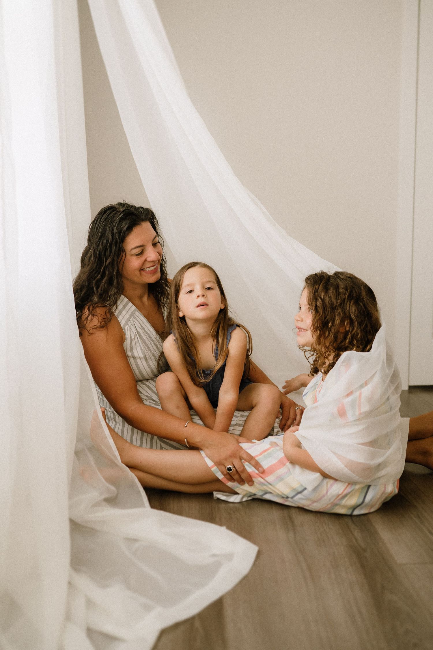 Family enjoys playful bonding time together sitting on floor with flowing white fabric backdrop.