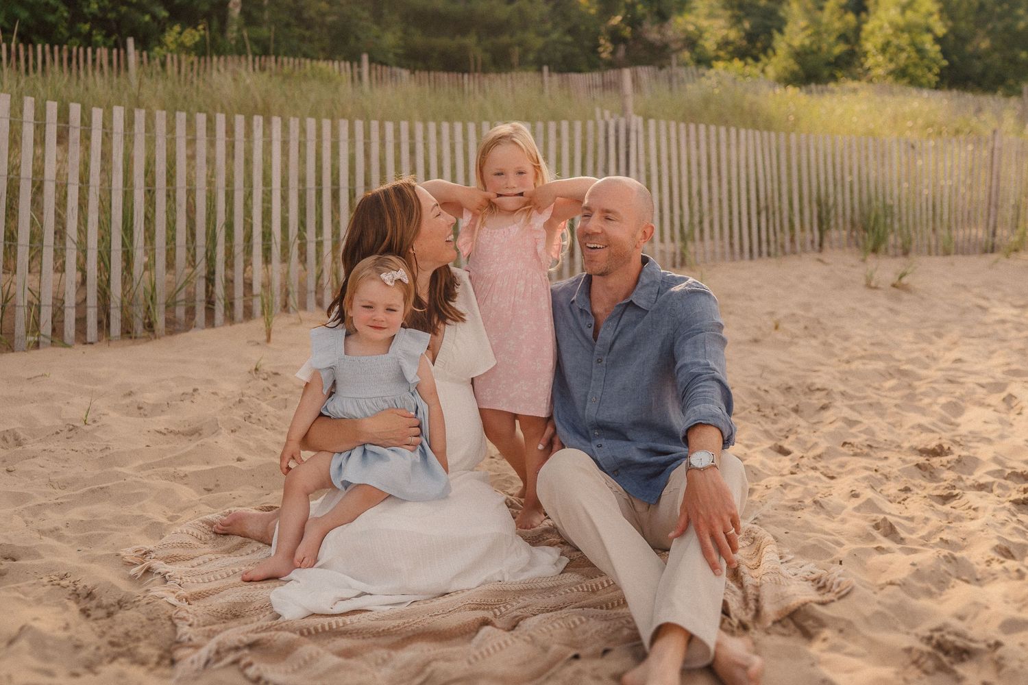 A family poses together on a sandy beach with wooden fencing and green foliage behind them.
