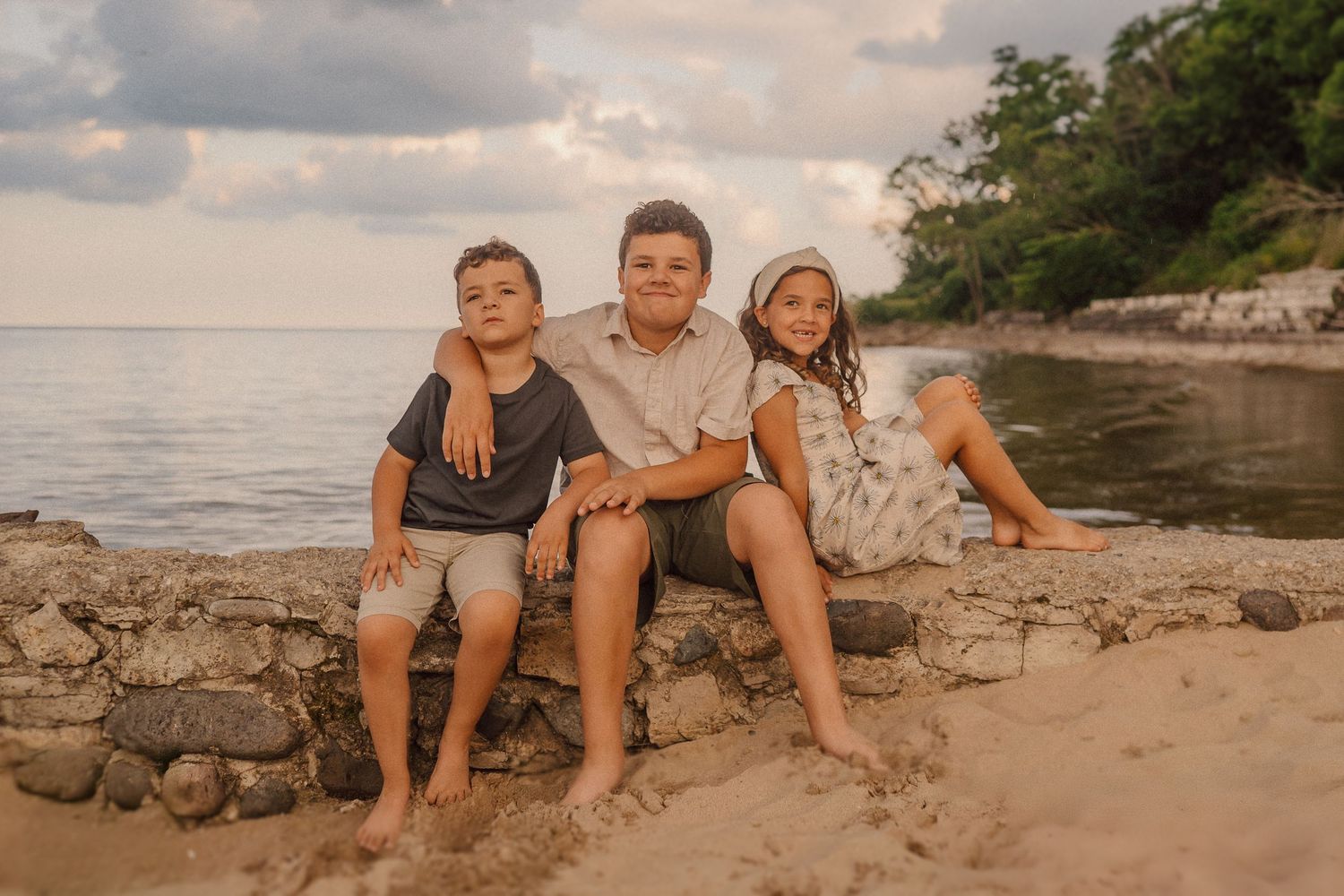 Three siblings sit together on a rocky beach shore at sunset with the ocean and trees in the background.