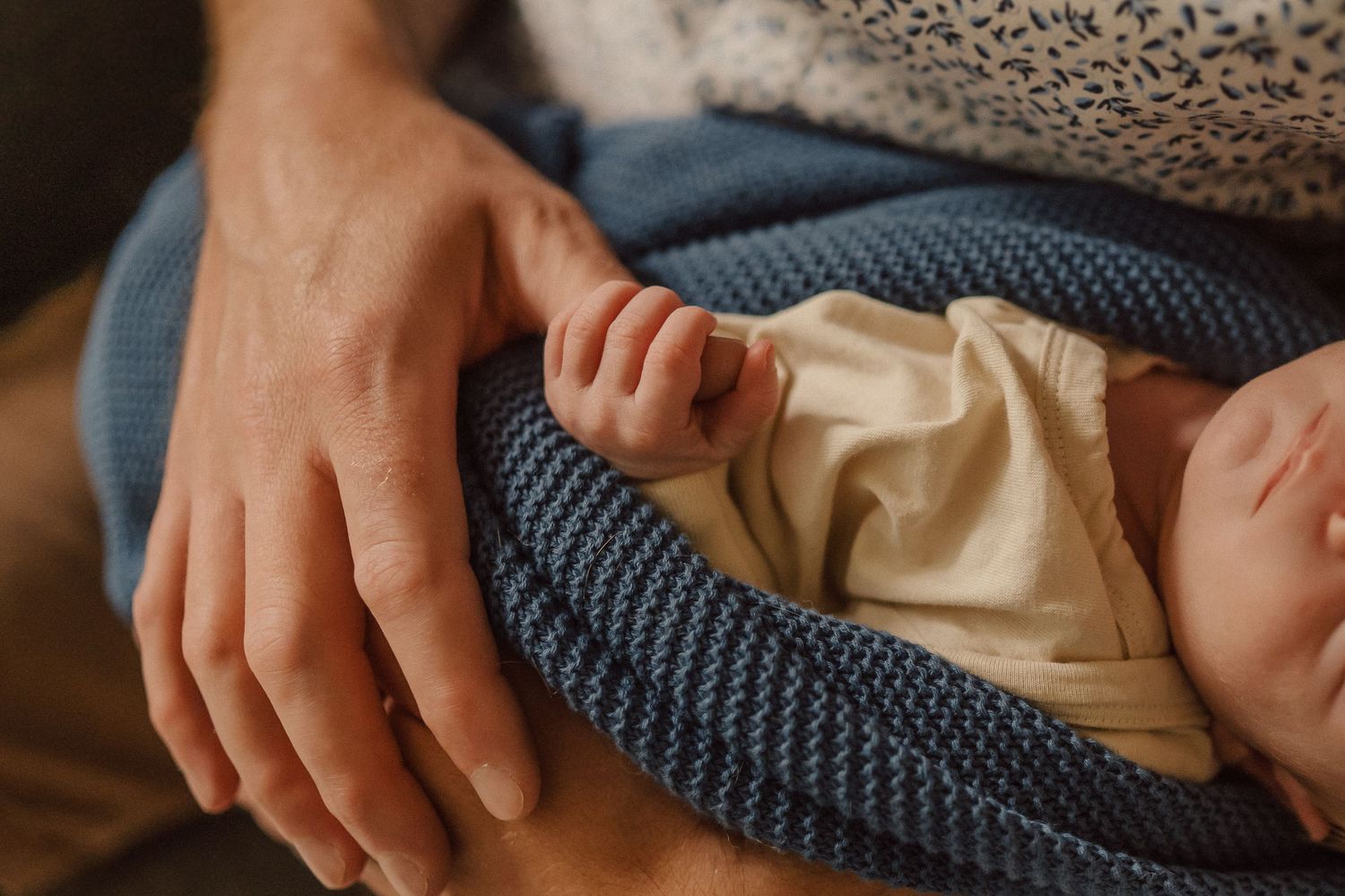 Close-up view of gentle hands cradling a sleeping newborn in cream and blue clothing.