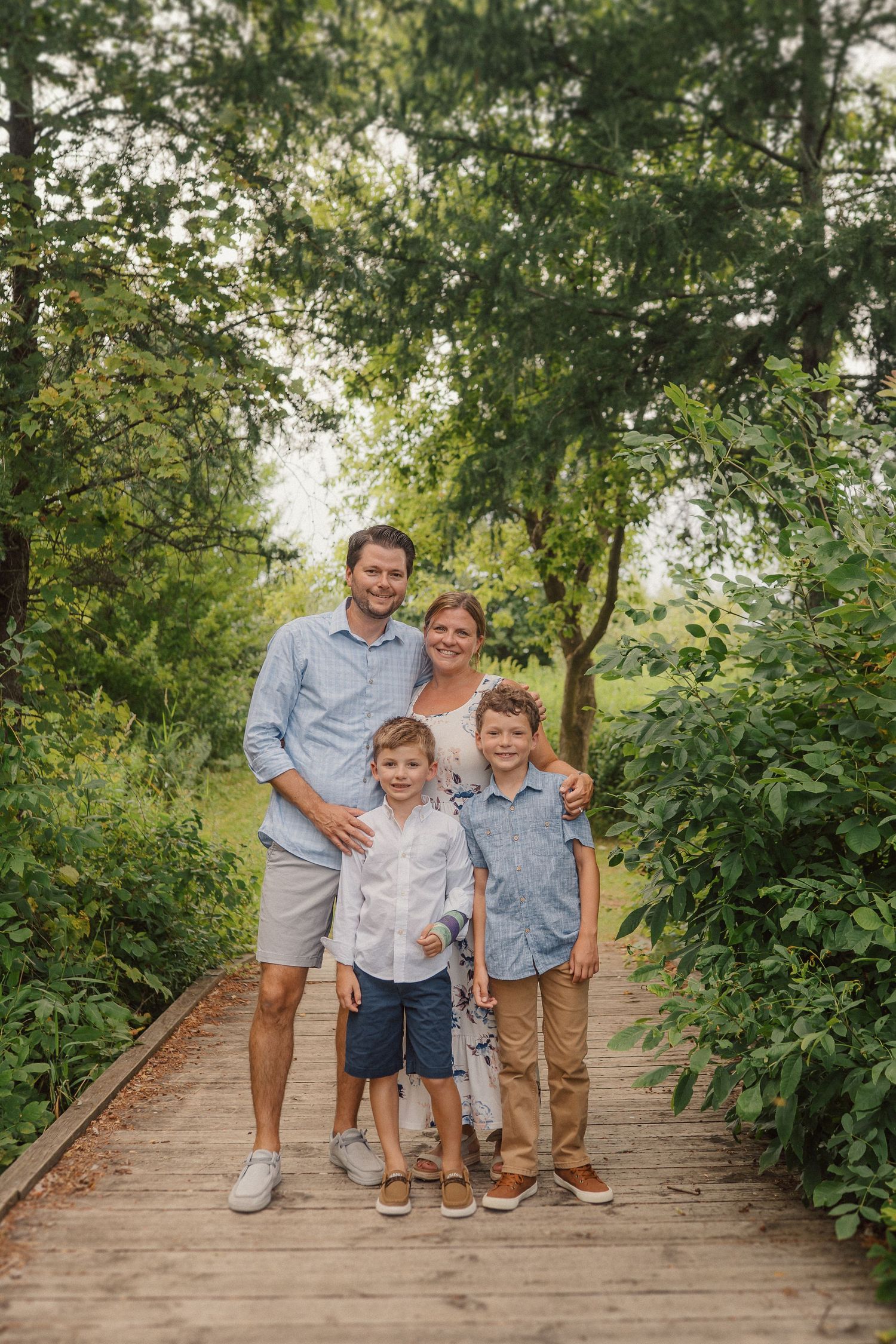 A family stands together on a wooden boardwalk surrounded by lush greenery on a sunny day.