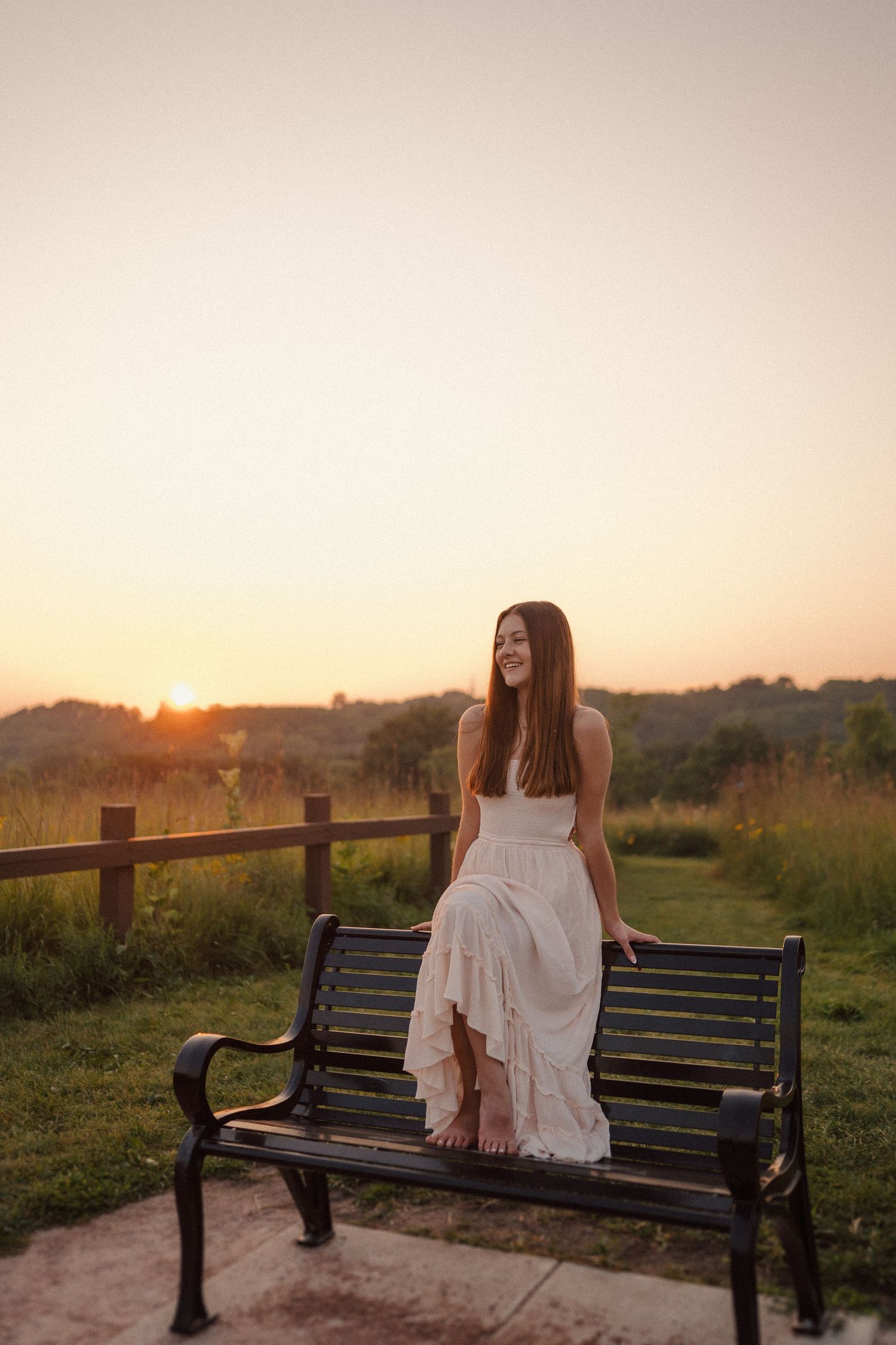 A woman in a white dress sits on a wooden bench during sunset in a scenic outdoor setting.