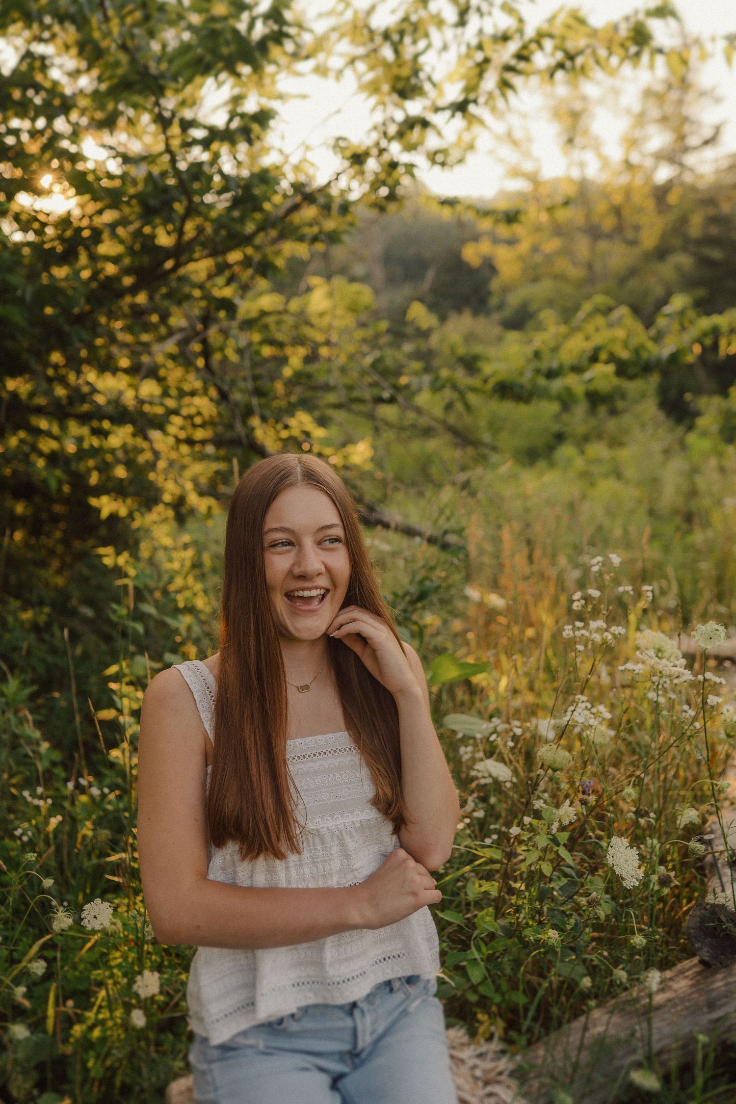 Person in white top and jeans sits amid wildflowers and sunlit foliage in a natural outdoor setting.