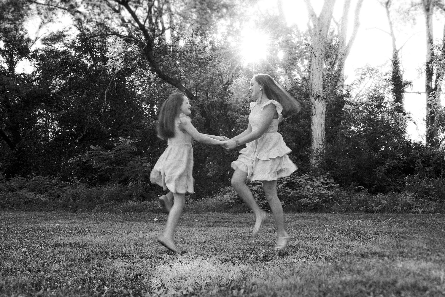 Two people in white dresses dance and twirl together in a backlit forest setting.