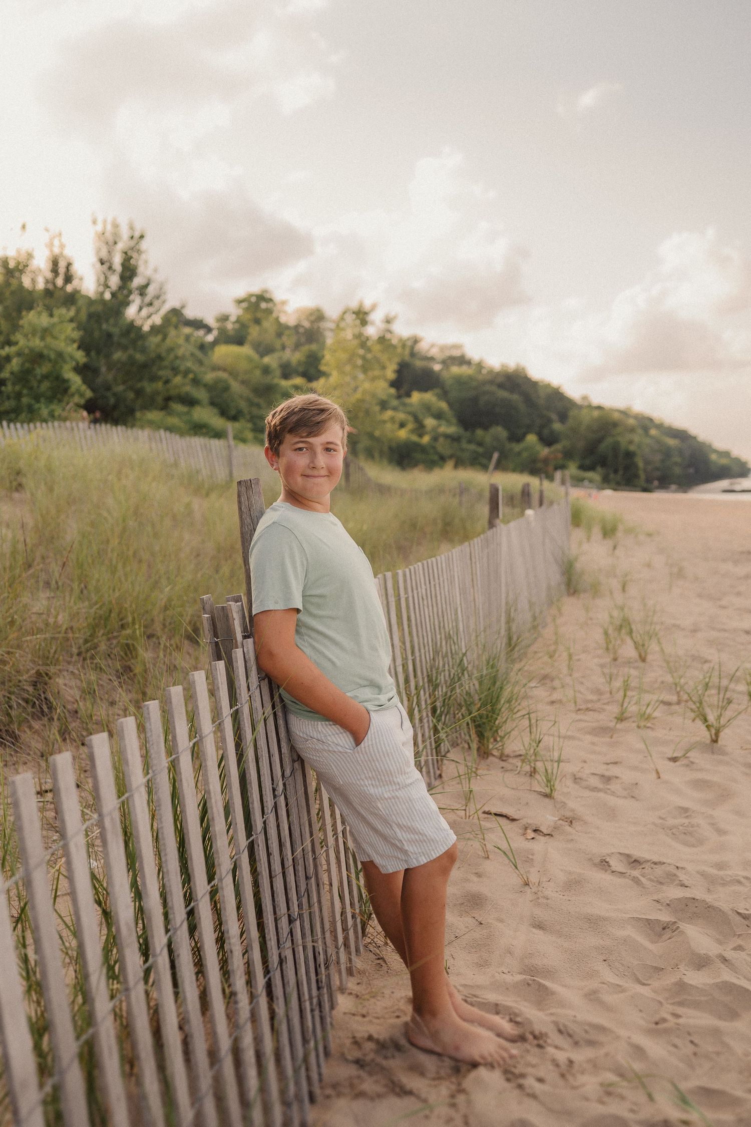 A photograph of someone standing barefoot on a sandy beach next to a wooden fence during sunset.