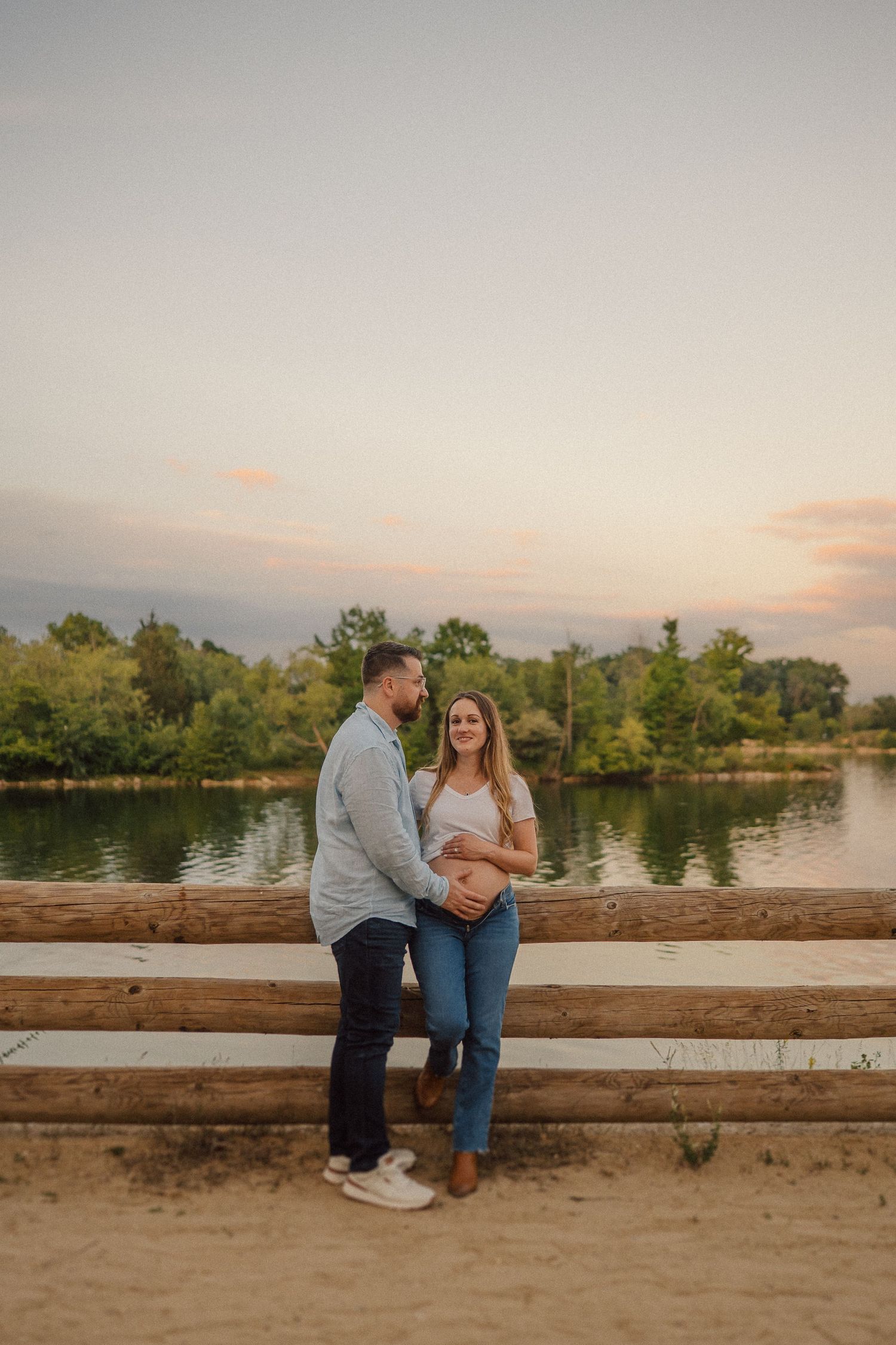 Couple stands on wooden dock by serene lake during sunset, with trees and soft pink sky in background.