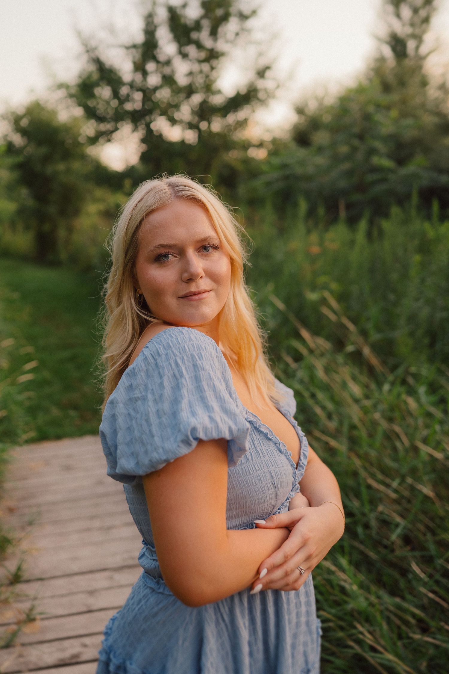 A person in a blue puff-sleeve dress poses on a wooden boardwalk surrounded by greenery at sunset.
