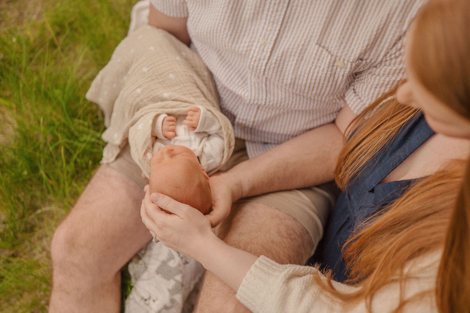 Outdoor family photo showing hands holding a sleeping baby on a sunny day in grassy setting.