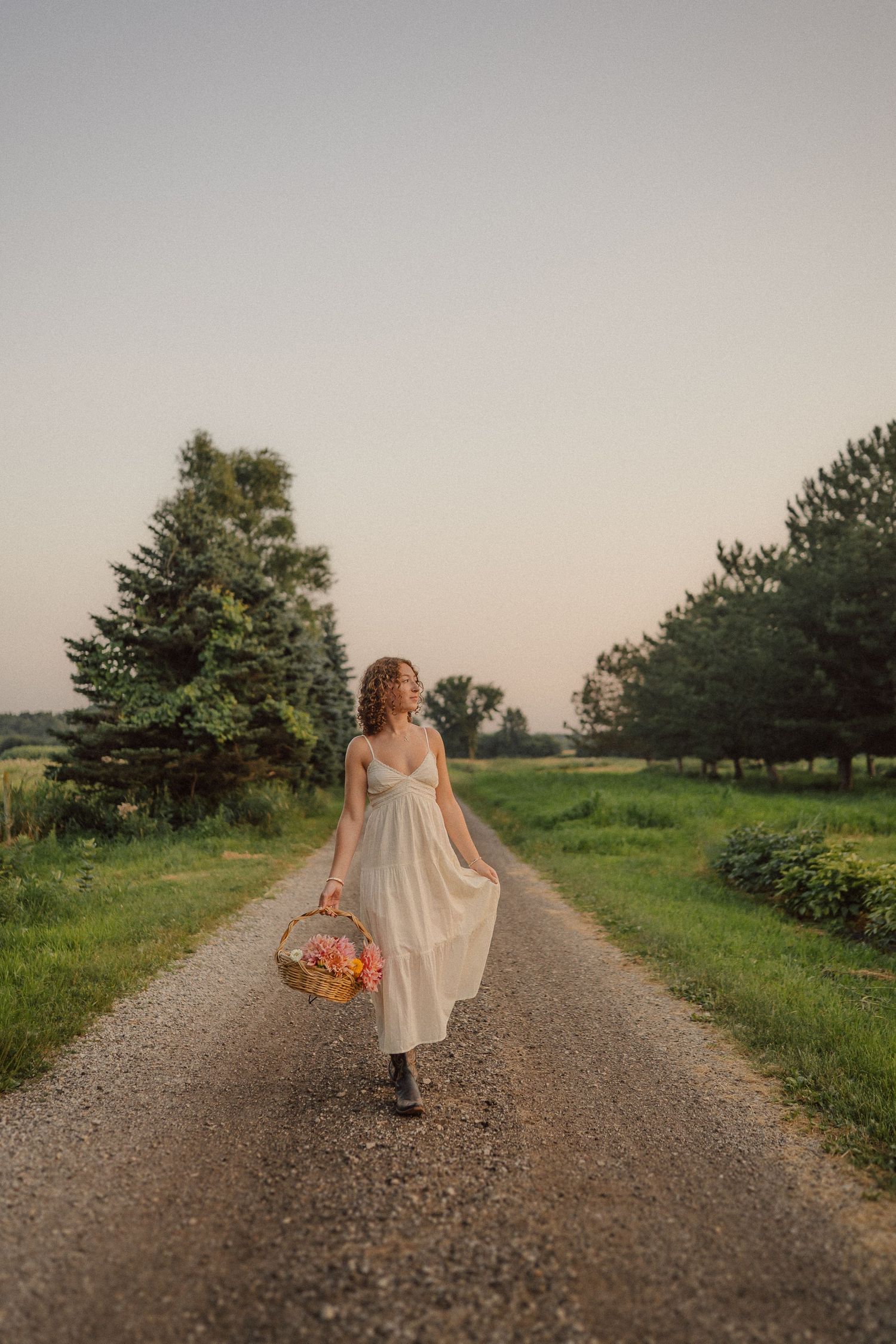 A person in a flowing dress walks along a gravel path through a green countryside at sunset with bright flowers in hand.