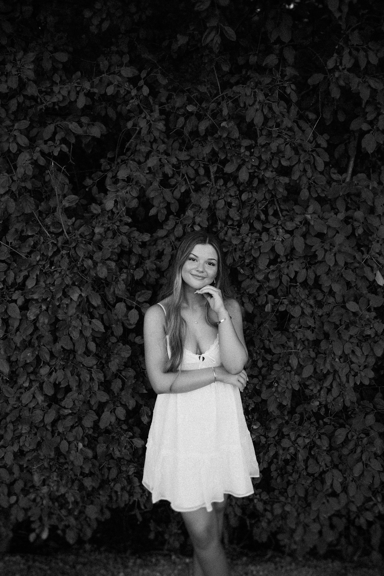 Black and white portrait of a woman in a white dress standing against a leafy wall backdrop.