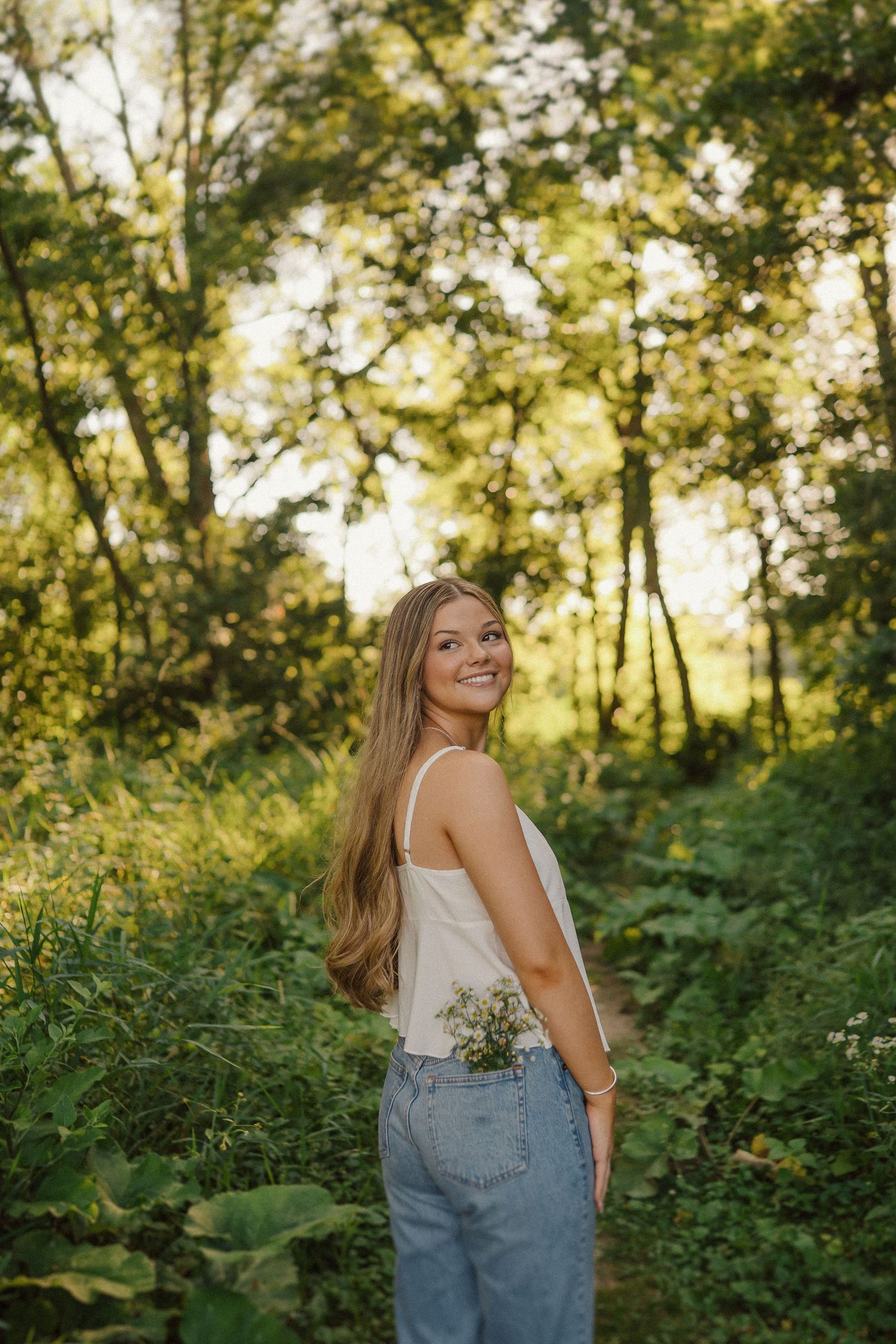 Person in overalls poses in a sunlit forest setting with greenery and golden light filtering through trees.