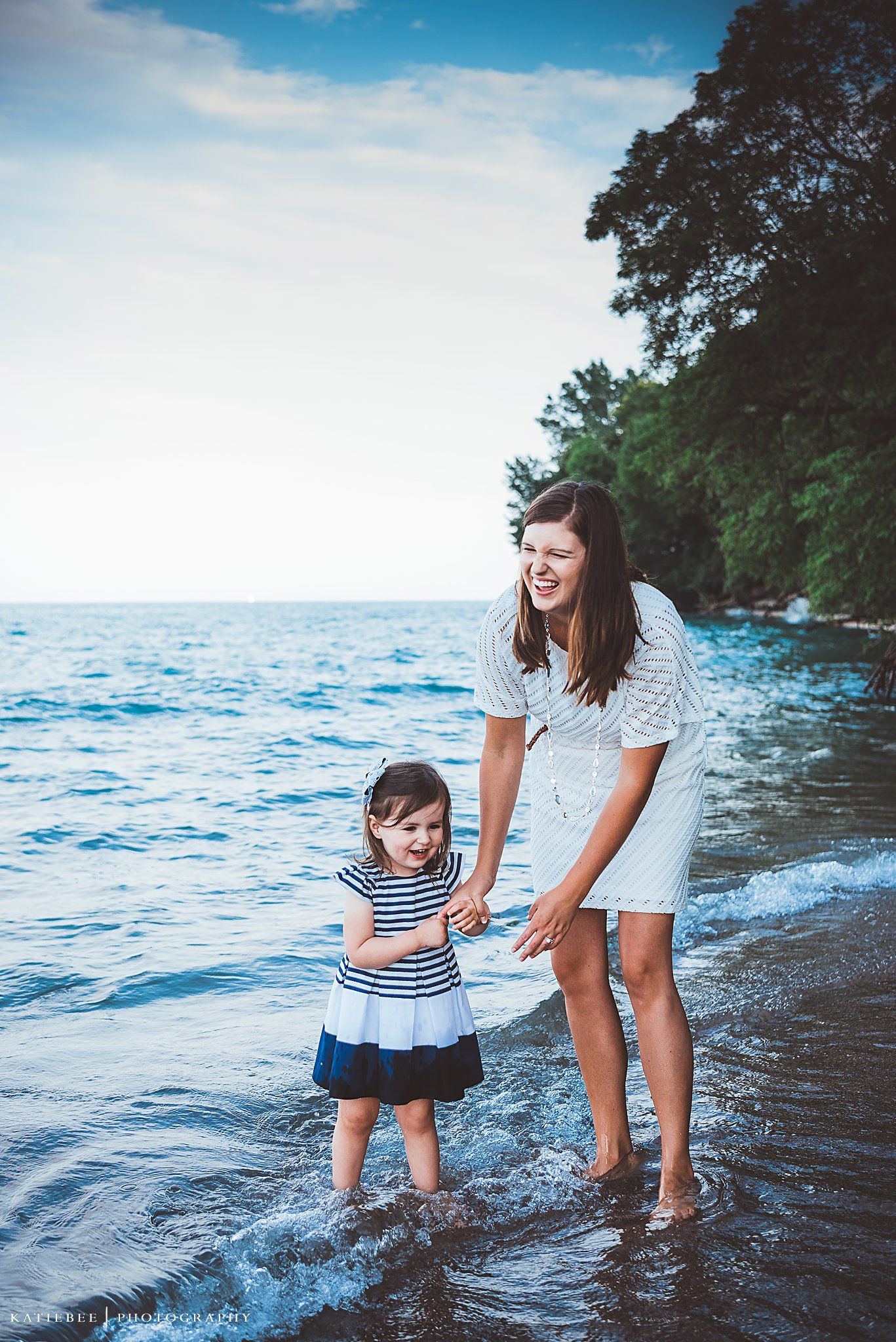 A parent and child play together in ocean waves at sunset near a wooded coastline.