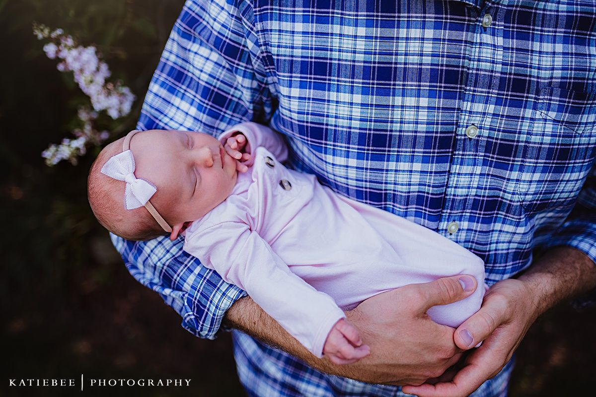 A tiny infant in white clothing rests peacefully against a blue plaid shirt in an outdoor portrait setting.