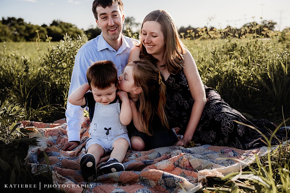 Family of four sitting together on a patterned blanket in a sun-drenched field.