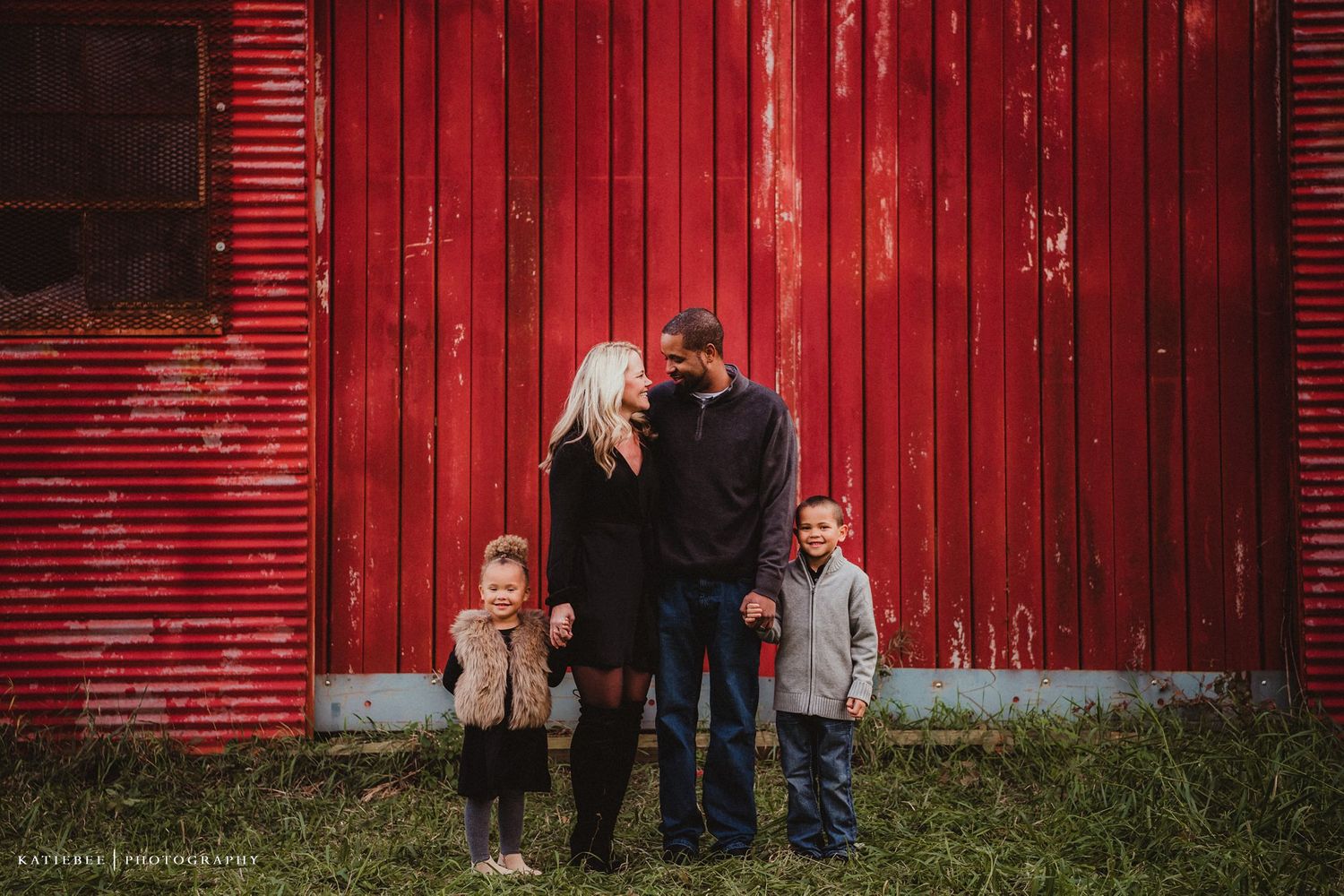 Family portrait against vibrant red barn wall showing authentic connection and joy.