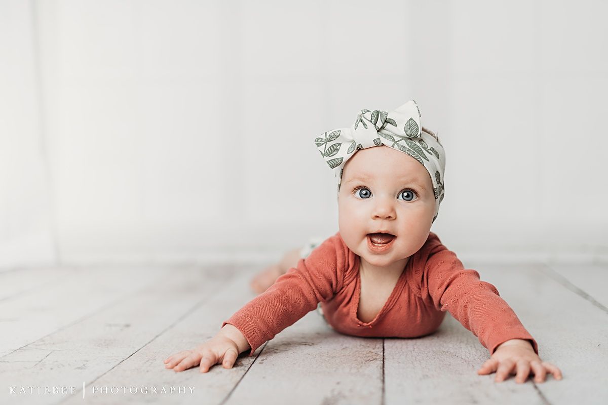 Smiling baby wearing coral top and floral headband on white wooden floor.