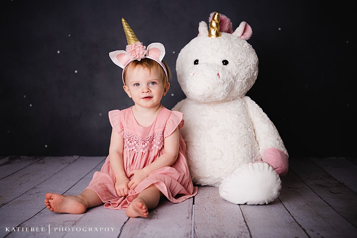 A pink-dressed toddler wearing unicorn ears sits next to a large plush white unicorn against a dark backdrop.