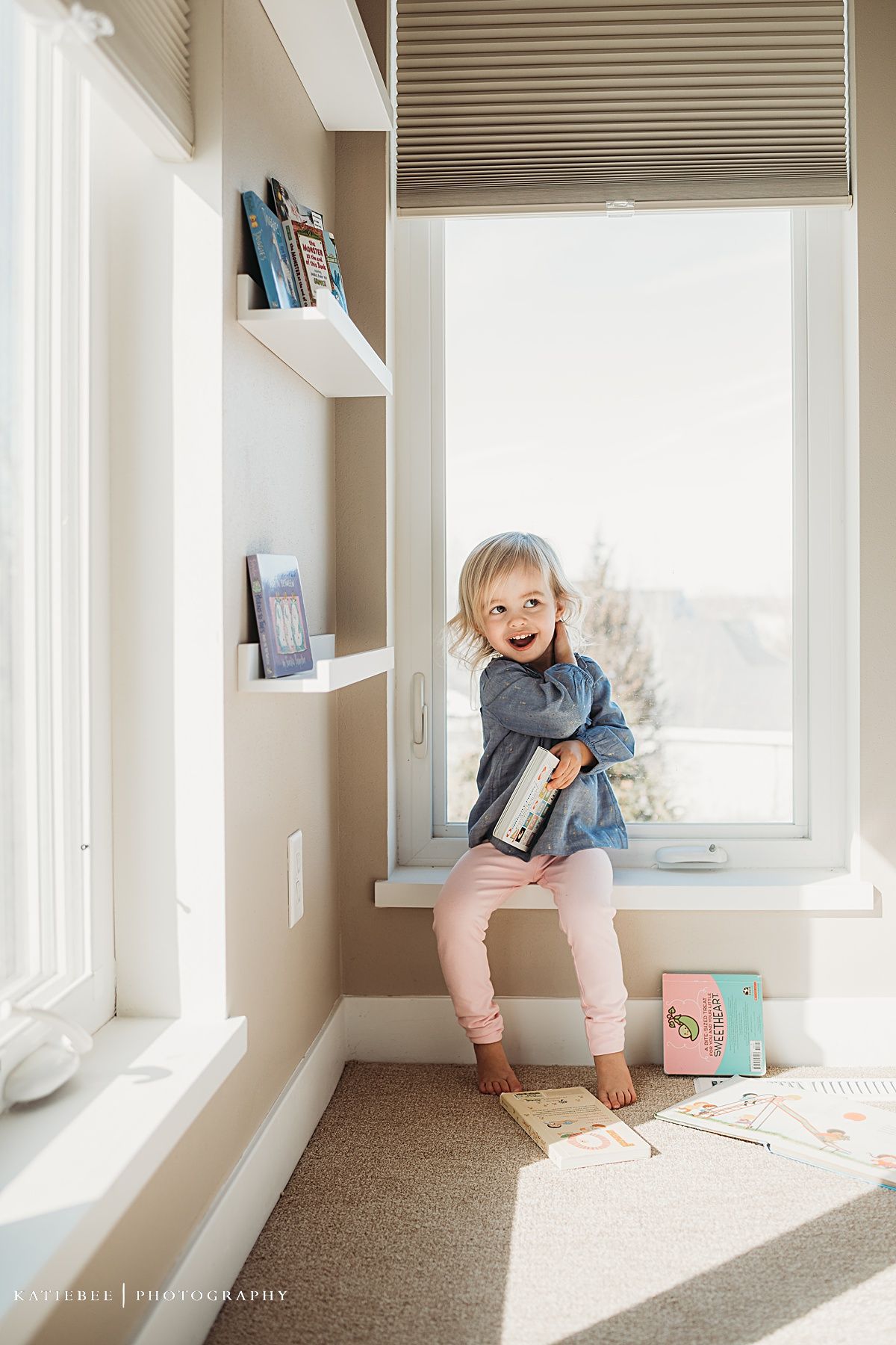 A toddler sits in a sunny window nook with white shelving and natural light streaming in.