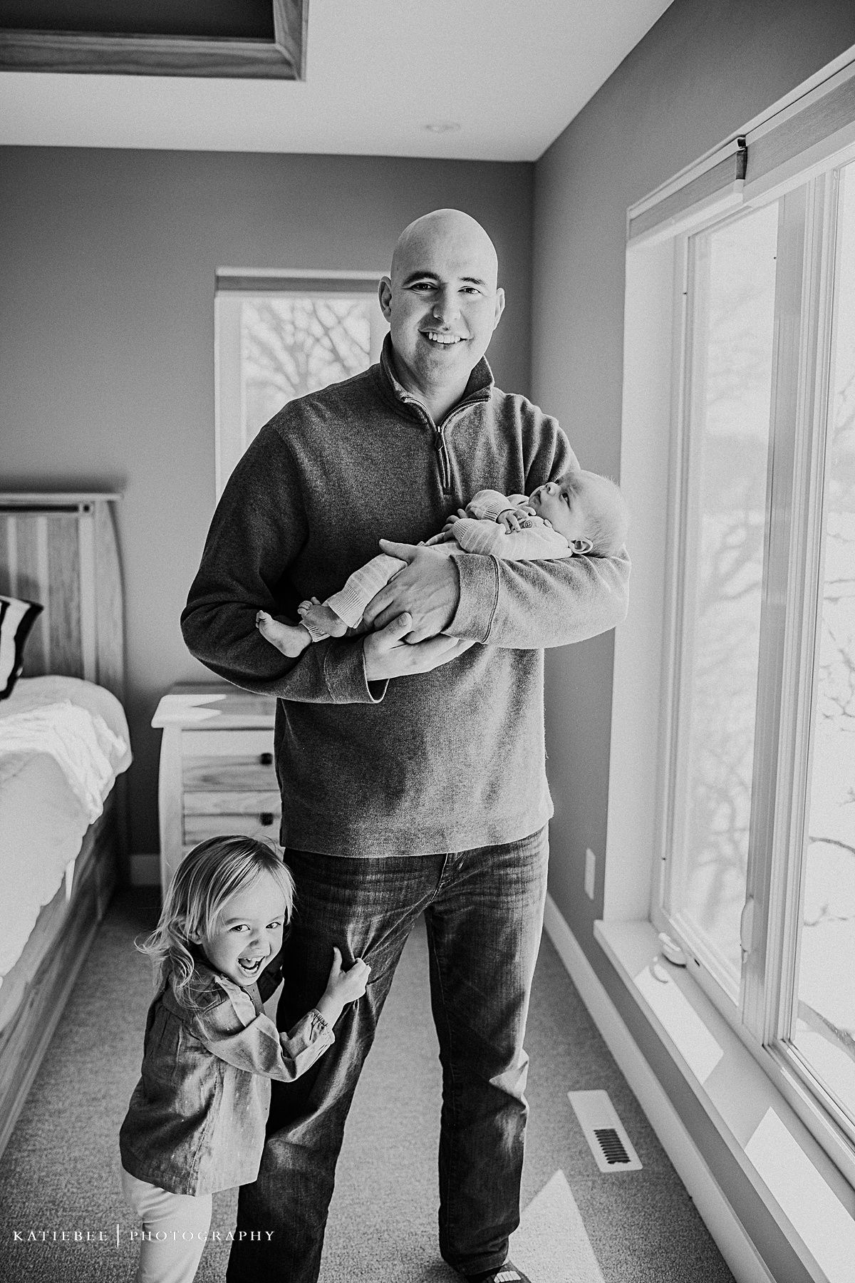A person in a grey sweater holds a newborn baby while standing in a sunny bedroom.