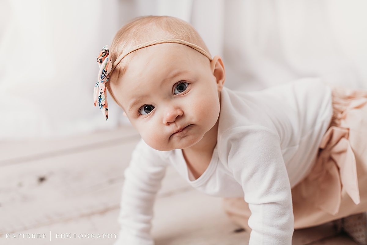 Baby in white onesie with floral headband crawling on light wooden floor.