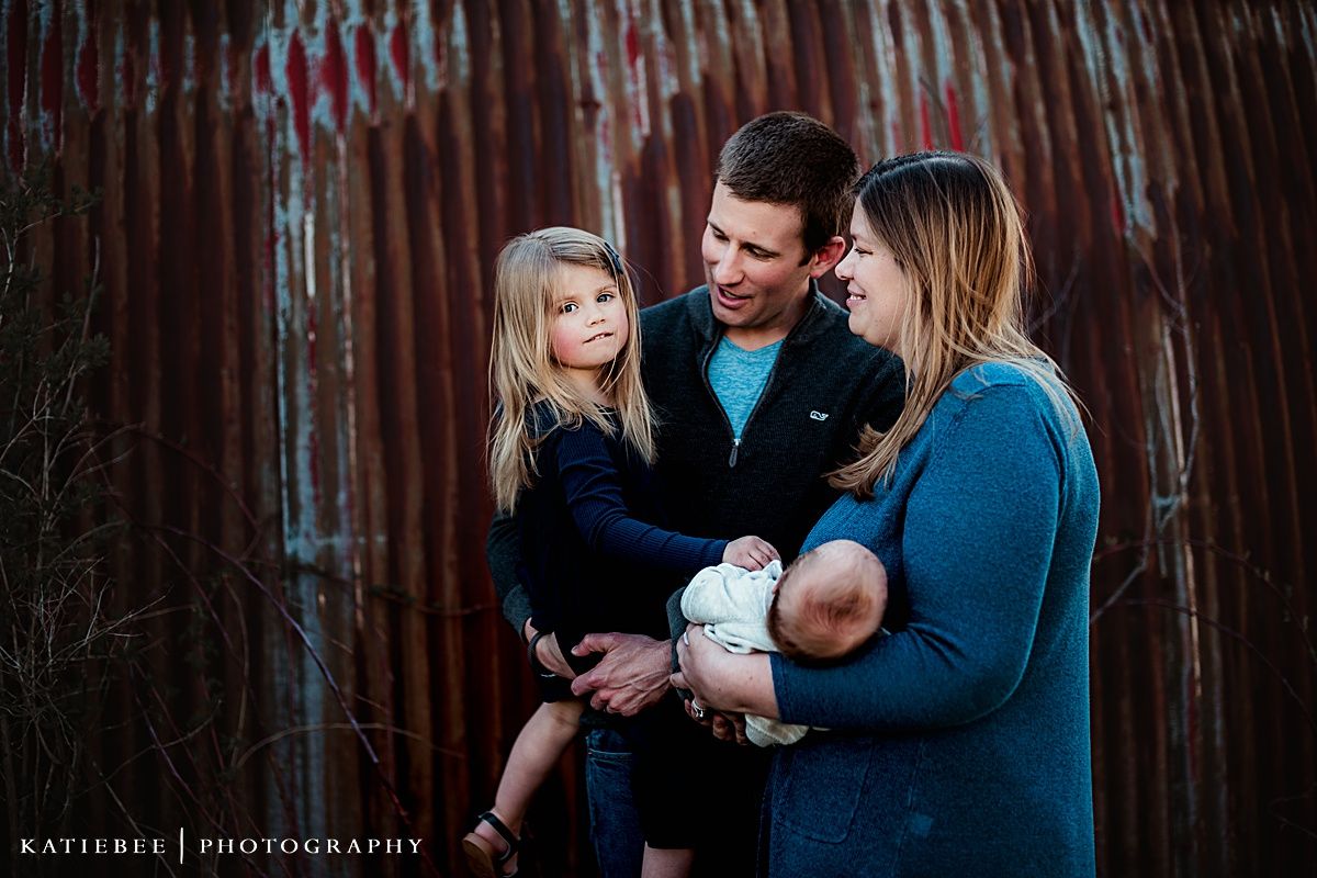 Family portrait against rustic metal wall in soft lighting.