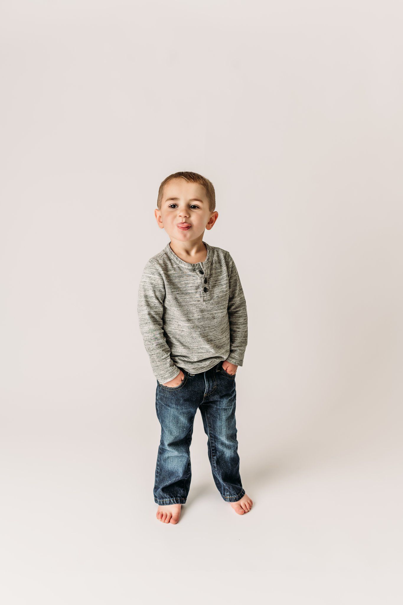 Standing pose with hands in pockets wearing gray sweater and jeans on white background.