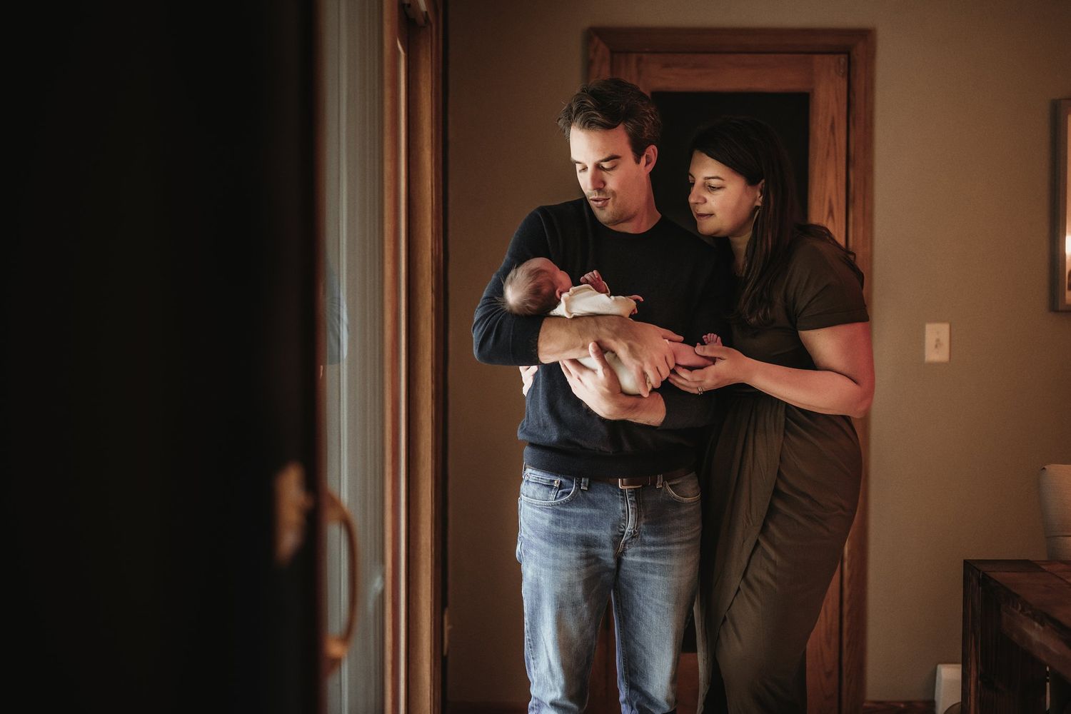 A couple stands in a doorway holding their newborn baby together in a tender moment.