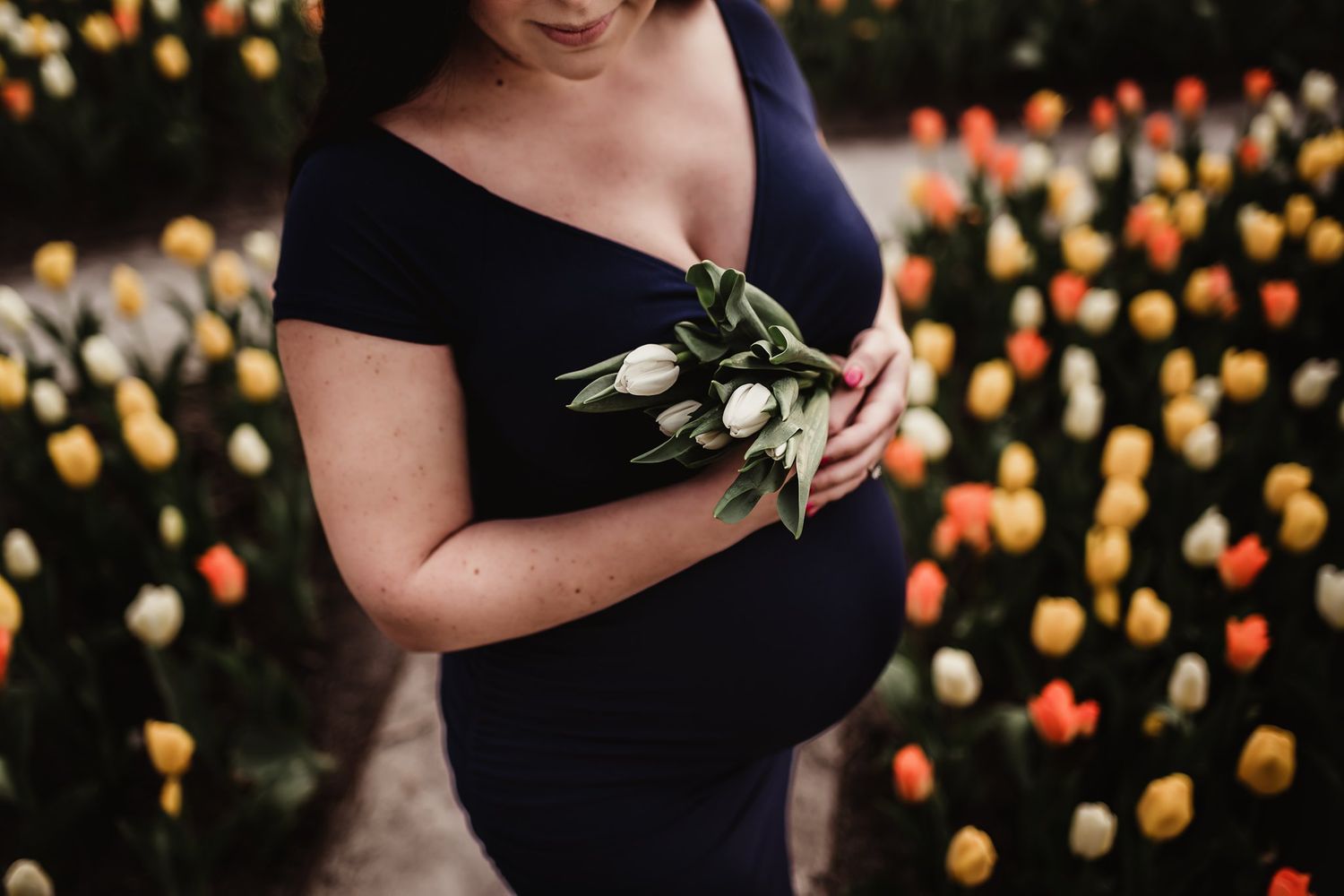 Close-up of pregnant belly in navy dress with olive branch against tulip garden.