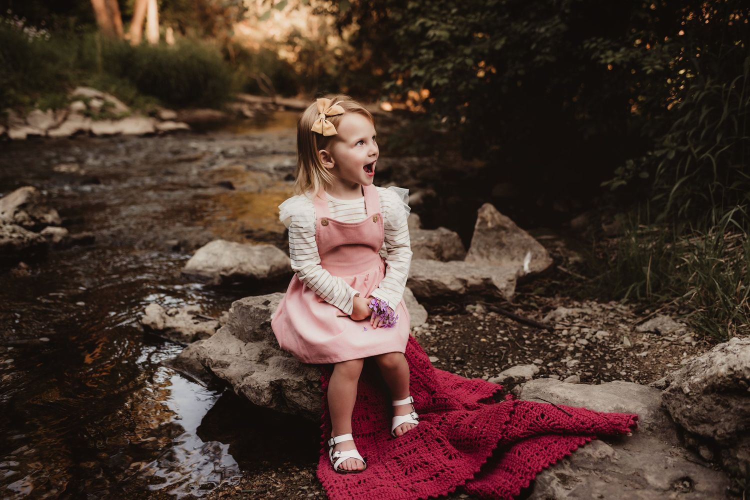 A child in pink dress and cream sweater sits on red blanket by stream.