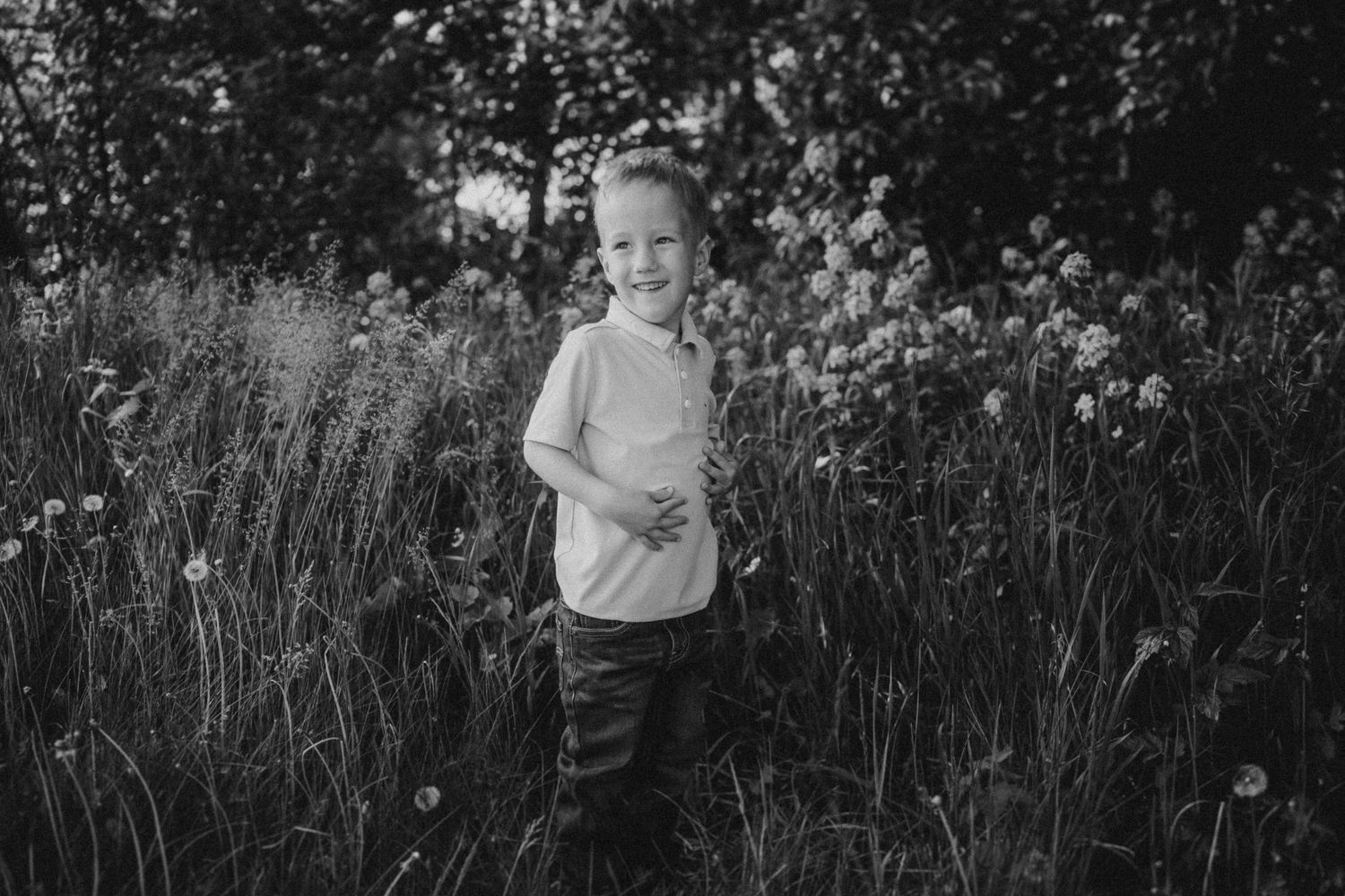 A young child stands in a field of tall grass and wildflowers in a black and white portrait.