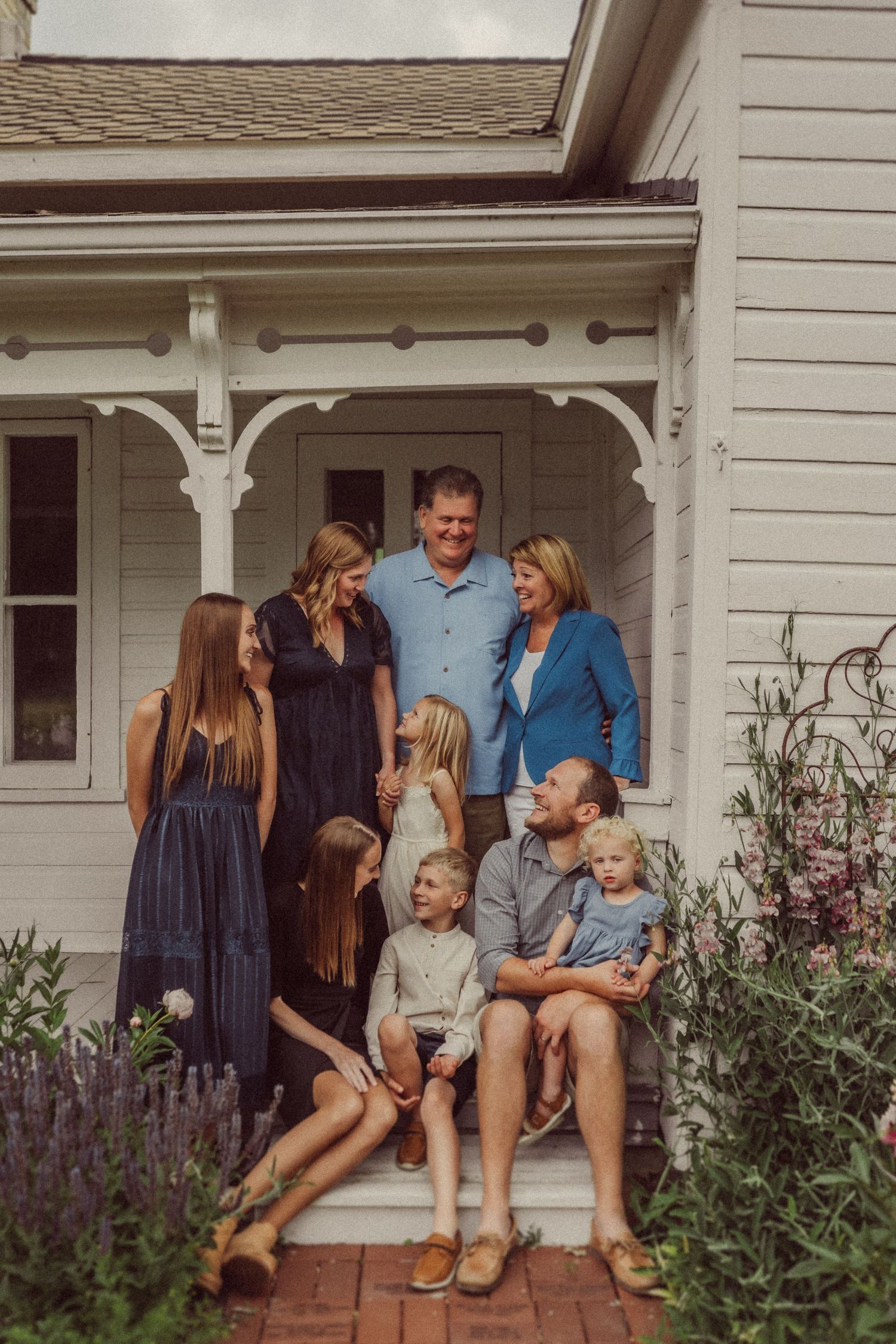 A family gathers on the front steps of their white farmhouse for a portrait among the garden flowers.