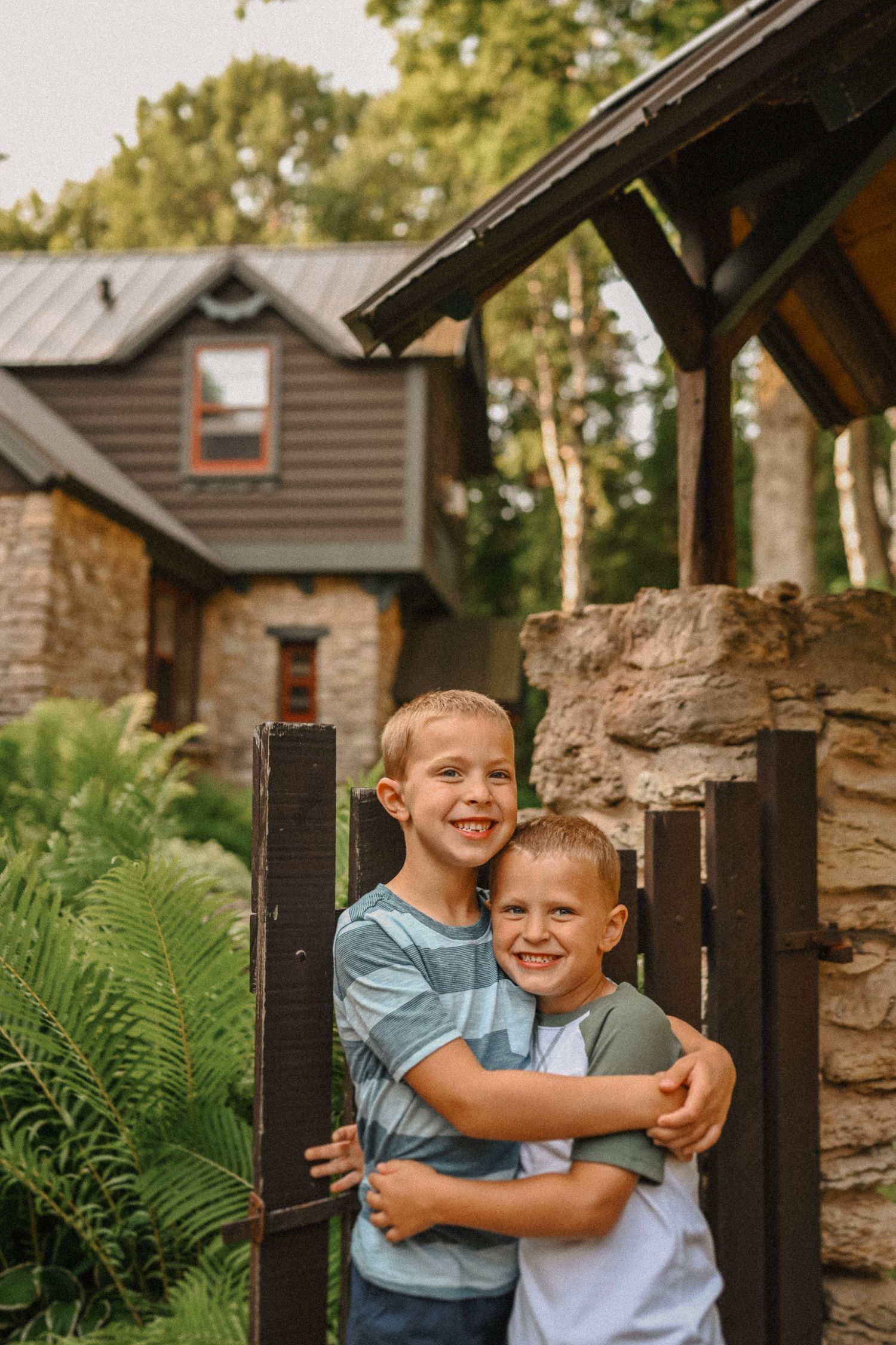 Two young boys in casual clothes share warm hug by rustic cabin gate.