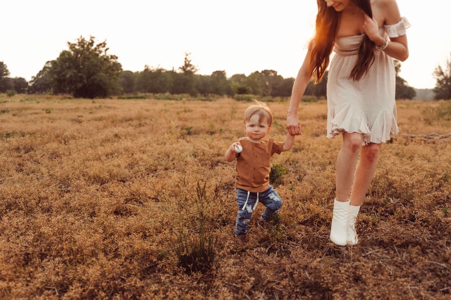 A small child walks through a sun-drenched field while holding hands with someone.