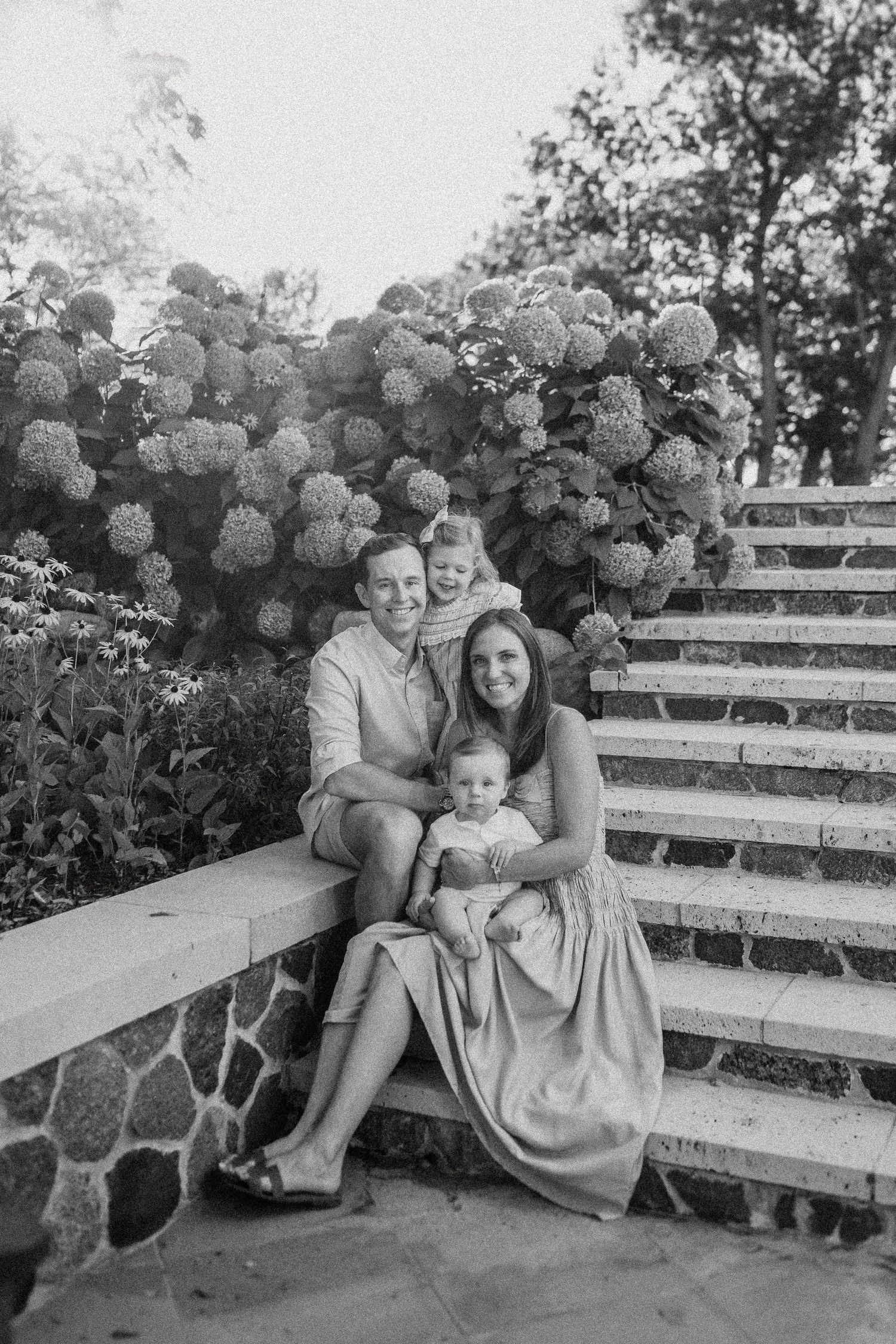 Black and white portrait of a family sitting on stone steps with lush hydrangea bushes in the background.