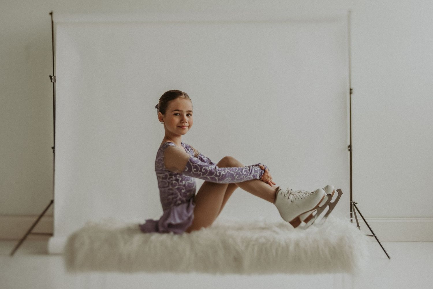 Ice skater in purple lace dress poses elegantly on white fur surface in studio setting.