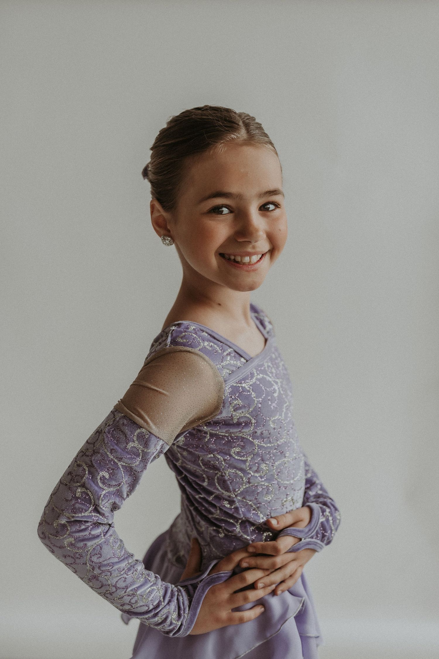Figure skater smiles in purple performance dress with intricate pattern against white background.