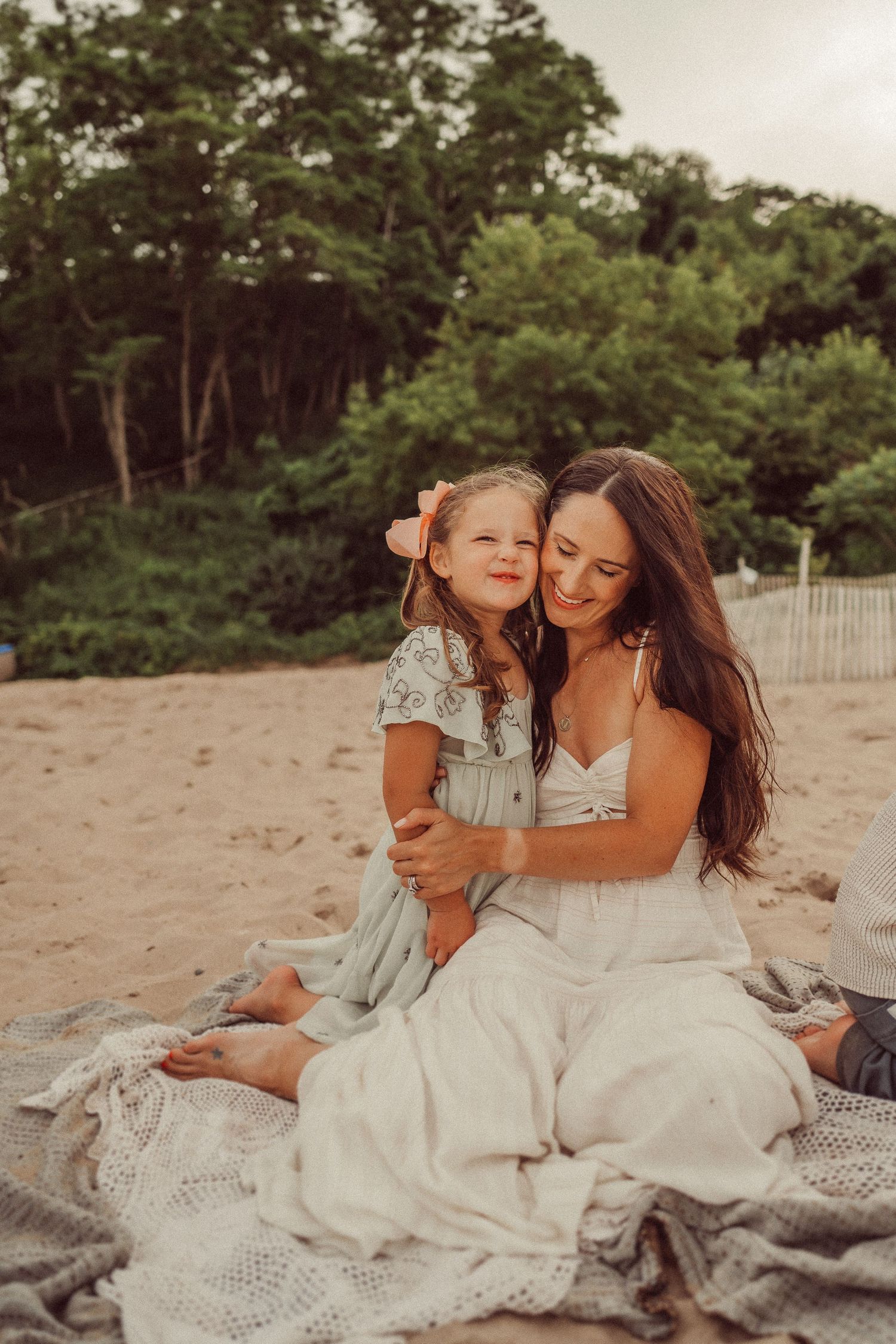 Two people in light colored dresses share happy smiles while sitting on a beach blanket.
