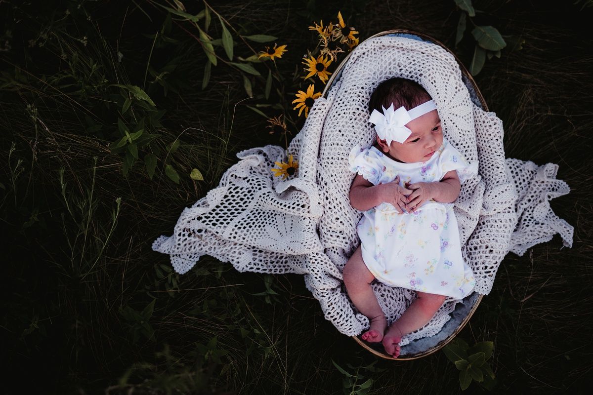 A baby in white dress and bow rests on intricate lace blanket surrounded by yellow wildflowers.