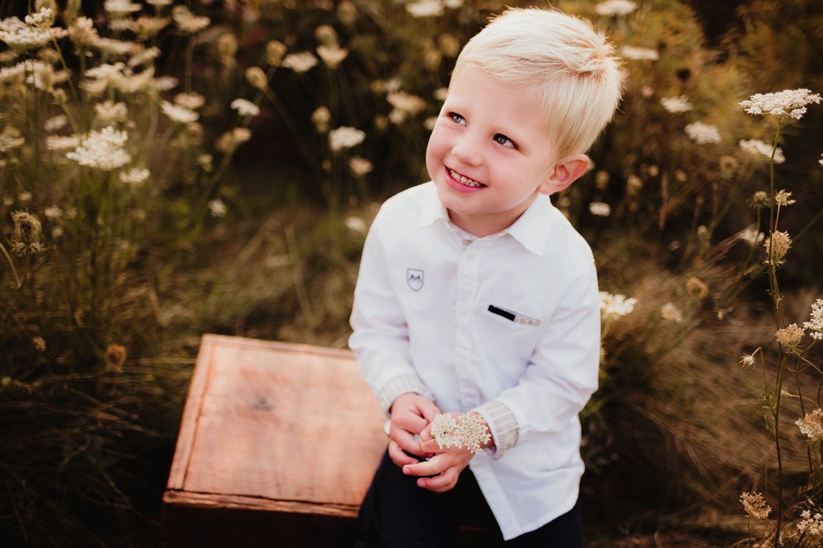 Child in crisp white button-down shirt sitting by wooden surface outdoors among wildflowers.