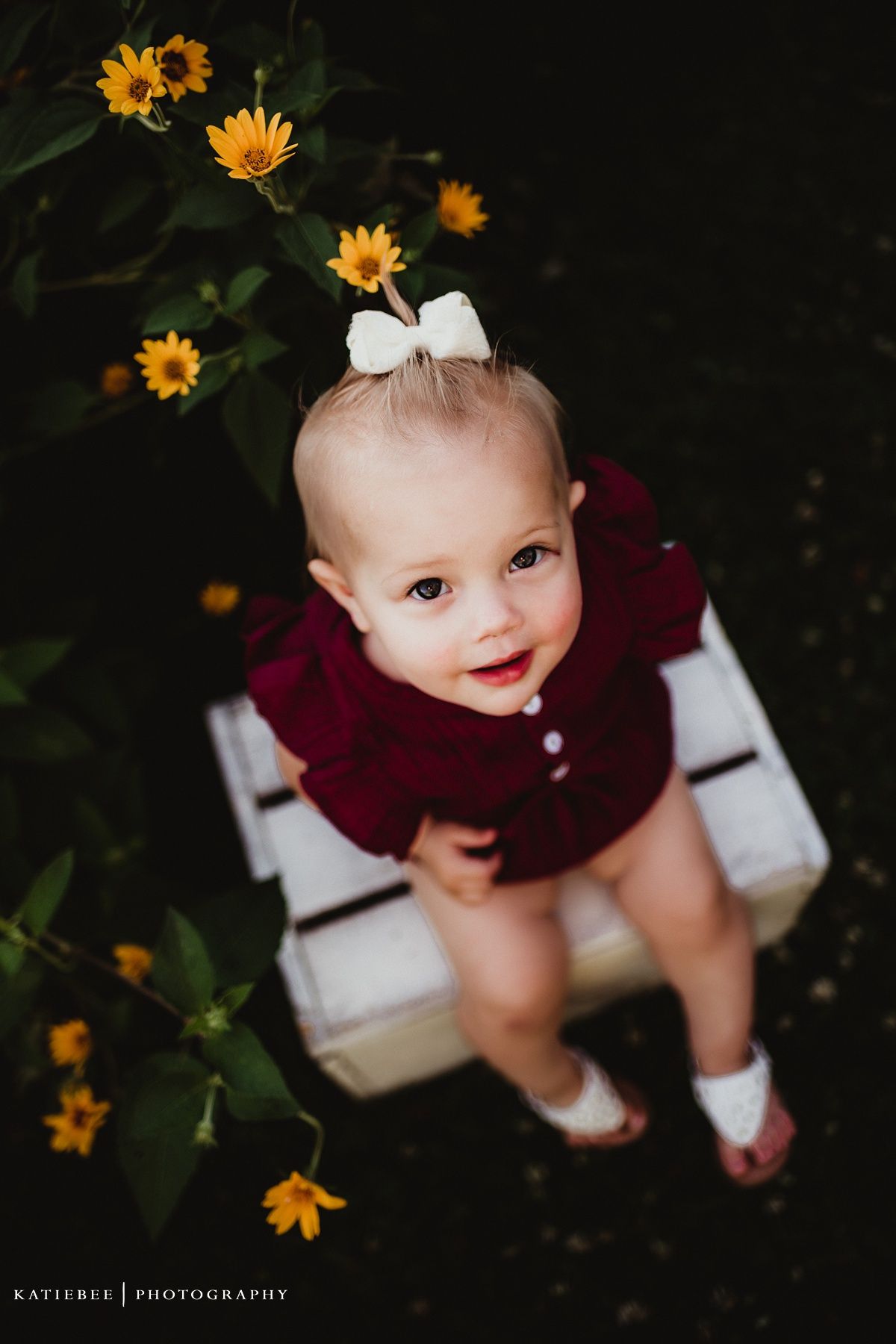 A small figure in a burgundy sweater sits on a white bench surrounded by yellow flowers.